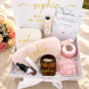 May include: A gift box with the name "Sophia" in gold script contains a variety of items. These include a sleep mask, a small cosmetic bag, a candle, a lip balm, a bottle of essential oil, a bath bomb, and a decorative soap. A calendar card reads "Will you be my bridesmaid?"