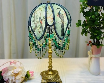 Puede incluir: Una lámpara de mesa decorativa con un diseño de pavo real en la pantalla. La pantalla es de color verde claro con un ribete azul oscuro y flecos de cuentas. La base es dorada y ornamentada. La lámpara está sobre una superficie blanca.