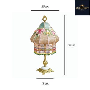 May include: A table lamp with a gold-toned metal base, a white circular accent, and a fabric shade featuring floral embroidery in pink, yellow, and green. The shade has a beaded fringe and measures 30 cm wide by 60 cm high, with a 15 cm base.