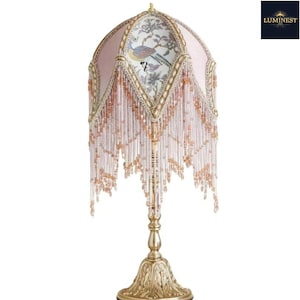 May include: A table lamp with a gold-toned metal base and a dome-shaped shade featuring pink fabric panels, floral and bird illustrations, and long, dangling light pink and clear glass beads. The word LUMINEST is printed in the top right corner.