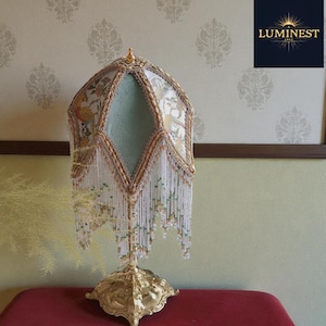 May include: An ornate, antique-style lamp with a gold-colored base and a decorative shade. The shade features a floral pattern with gold trim, a mint green center, and beaded fringe. The lamp is on a red surface.