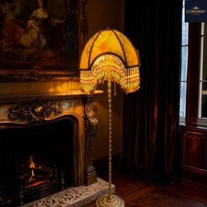 May include: A vintage-style floor lamp with a yellow lampshade adorned with a floral pattern and beaded fringe. The lamp has a gold-colored metal base and stem. The lamp is placed near a fireplace and a window.