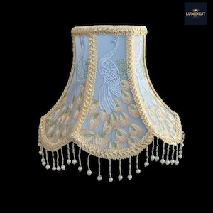 May include: A light blue lampshade with embroidered peacock designs in white, green, and peach. The shade is trimmed with gold-colored braid and dangling beaded accents, creating an elegant, decorative lighting element.