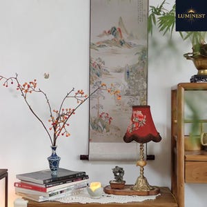 May include: A decorative table lamp with a red floral lampshade and a brass base. A vase with branches and berries sits on a stack of books. A traditional landscape painting hangs on the wall, creating a calm atmosphere.