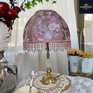 May include: A vintage table lamp with a pink lampshade adorned with floral embroidery and beaded fringe. The lamp has a gold base and a white stem. The lamp is on a table with a lace doily.
