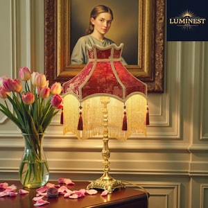 May include: A decorative table lamp featuring a red floral patterned shade, a gold base, and tassel detailing. The lamp is set on a dark wooden surface, accompanied by a vase of pink tulips and a framed portrait. The brand logo "LUMINNEST 1865" is visible.