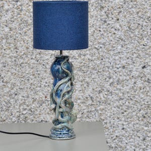 Könnte beinhalten: Eine Keramik-Tischlampe mit einem marineblauen Stoffschirm. Der Lampenfuß ist ein blau-grünes Oktopus-Design. Die Lampe steht auf einer grauen Oberfläche.