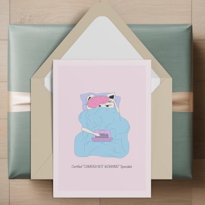 Op de afbeelding: Een lichtroze wenskaart met een cartoonkat onder een blauwe deken met een roze slaapmasker, typend op een kleine paarse laptop. De tekst onder de illustratie luidt: Certified "CAMERA NOT WORKING" Specialist. De kaart zit in een envelop.