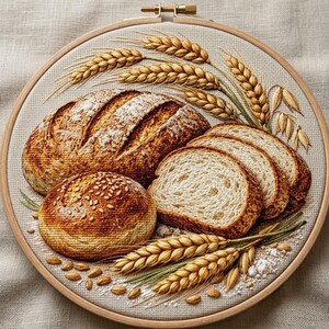 Puede incluir: Un bastidor de bordado circular que contiene un diseño detallado de un pan de masa madre grande, un panecillo redondo con semillas y dos rebanadas de pan, todo rodeado de espigas de trigo doradas sobre un fondo de tela beige claro.