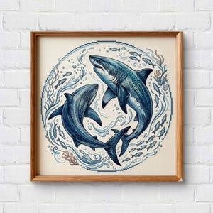 May include: A square wooden frame containing a circular cross-stitch embroidery featuring two blue sharks swimming in a clockwise motion. The design includes small fish, bubbles, and coral accents on a light beige fabric background.