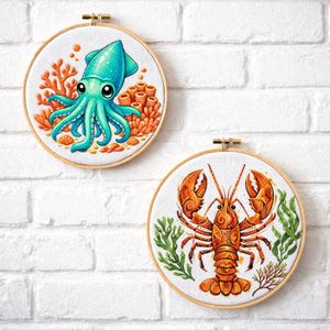 May include: Two circular embroidery hoops featuring cross-stitch designs. The top hoop displays a teal squid surrounded by orange coral. The bottom hoop shows an orange lobster with intricate patterns, flanked by green seaweed on a white fabric.