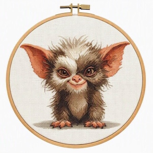 May include: A circular wooden embroidery hoop containing a cross-stitch pattern of a small, furry creature with large, pointed ears, brown and white fur, and dark eyes. The design is centered on a white fabric background within the frame.