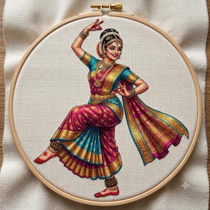 May include: A cross-stitch embroidery in a circular wooden hoop featuring a dancer in a vibrant magenta and teal sari with gold accents. The figure is posed with one leg lifted, wearing traditional jewelry, headpiece, and ankle bells on white fabric.