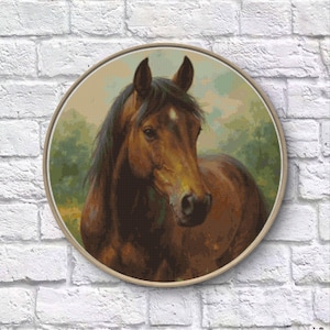 May include: A circular cross-stitch depicting a brown horse's head, framed in light wood. The horse has a dark mane and a white star on its forehead. The background shows a blurred landscape with green and yellow tones. The artwork is displayed against a white brick wall.