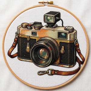 May include: A cross-stitch embroidery of a vintage-style camera with a black and tan body, a large central lens, and a top-mounted flash unit. The camera is framed in a circular wooden hoop against a white fabric background with a brown leather strap.