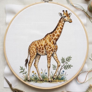 May include: A cross-stitch embroidery project in a round wooden hoop featuring a giraffe with brown and tan spots standing among small green plants and yellow and blue flowers on white Aida fabric, accompanied by metal scissors and thread spools.