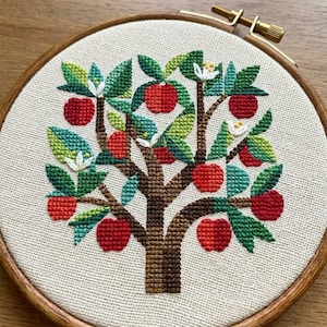 以下が含まれることがあります： 円形の木製刺繍枠に入ったクロスステッチ刺繍。茶色の幹、緑の葉、白い花、赤いリンゴが描かれた木のデザインです。織り目の見える明るいベージュの布地に刺繍が施されています。