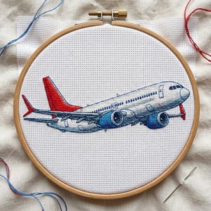 Könnte beinhalten: Ein Kreuzstich-Stickprojekt, das ein weißes Passagierflugzeug mit rotem Leitwerk und roten Flügelspitzen zeigt, eingespannt in einen runden Holzstickrahmen. Das Flugzeug ist mit blauem und rotem Garn auf weißem Aida-Stoff gestickt.