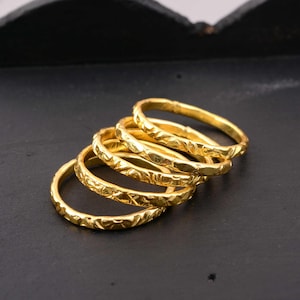 May include: A set of five gold-colored rings, each featuring a textured, hammered surface with a repeating cross-hatch pattern. The rings are stacked in a staggered, overlapping arrangement, showcasing a bright, metallic finish throughout.