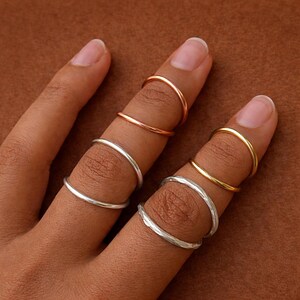 Può includere: Primo piano di una mano con diversi anelli alle dita. Gli anelli sono di vari colori, tra cui argento, oro e oro rosa. Gli anelli hanno un design semplice e minimalista. Lo sfondo è di colore marrone.