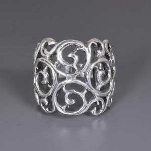 May include: A silver ring with an openwork design. The ring features a continuous pattern of swirling, scroll-like shapes. The metal has a polished finish, and the ring appears to be a wide band. The background is a solid grey.