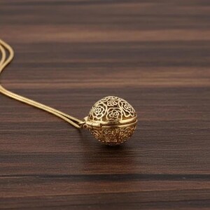 Puede incluir: Un collar dorado con un colgante esférico calado con un diseño floral. La fina cadena de oro conduce al colgante decorativo. El collar descansa sobre una superficie de madera marrón oscuro.