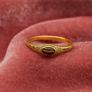 Könnte beinhalten: Ein goldener Ring mit einem ovalen, braunen Edelstein. Der Ring hat ein verziertes Design auf dem Band und um den Edelstein herum. Der Ring ist vor einem rosa Hintergrund platziert.