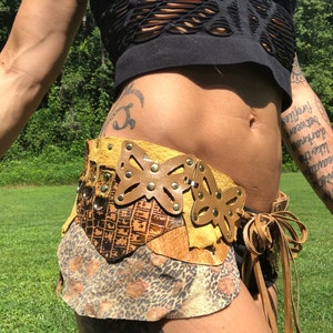 May include: A brown leather belt with a butterfly design and a leopard print fabric skirt. The belt has gold-colored metal accents and is tied with a brown leather cord.