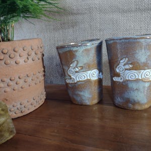 May include: Two ceramic tumblers featuring a hand-carved white rabbit design with swirling patterns. The cups have a mottled brown and blue-grey glaze finish. Each tumbler has a cylindrical shape with a slightly tapered base and a smooth rim.