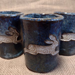 May include: Three cylindrical ceramic cups featuring a deep blue, mottled glaze with a glossy finish. Each cup displays a raised, white, relief-style illustration of a running hare with swirling fur patterns embossed on the exterior surface.