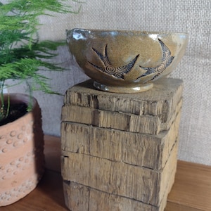 May include: A tan ceramic bowl with a glossy finish featuring two carved swallow silhouettes. The birds are decorated with intricate dark spiral patterns etched into the clay surface. The bowl has a rounded shape and a small circular base.