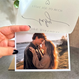 May include: A rectangular greeting card featuring a photograph of a couple embracing outdoors near water. The top flap is lifted to reveal text that reads I LOVE YOU SO MUCH, baby, and HAPPY BIRTHDAY, accompanied by a small heart icon.