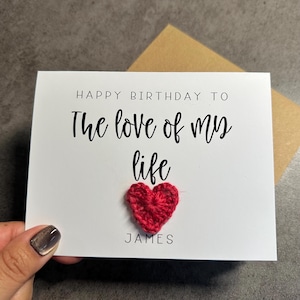 May include: A white rectangular greeting card featuring the black printed text "HAPPY BIRTHDAY TO The love of my life" above a small, textured, red crocheted heart, with the name "JAMES" printed in black text below the heart.