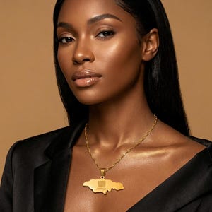Könnte beinhalten: Eine goldfarbene Halskette mit einem Anhänger in Form der Insel Jamaika. Der Anhänger hat ein Flaggen-Design und den Schriftzug "JAMAICA". Die Halskette wird von einer Person mit einem schwarzen Oberteil getragen. Die Kette ist gedreht.