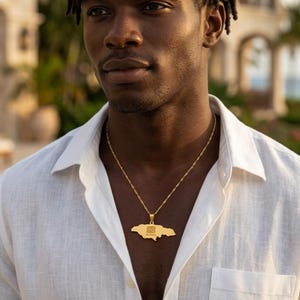 May include: A gold-colored pendant necklace featuring the outline of Jamaica. The pendant hangs from a delicate chain and is worn against a white button-down shirt. The necklace is a symbol of national pride.