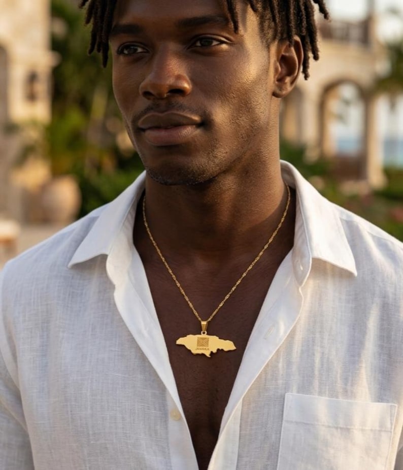 May include: A gold-colored pendant necklace featuring the outline of Jamaica. The pendant hangs from a delicate chain and is worn against a white button-down shirt. The necklace is a symbol of national pride.