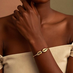 May include: A gold bracelet with two seashell-shaped charms. The bracelet is worn on a dark arm, with the charms on the wrist. The background is a soft blend of beige and green.