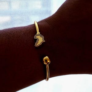Puede incluir: Brazalete abierto de color dorado. El brazalete presenta un dije con forma de continente, posiblemente África, con una superficie texturizada. El brazalete se lleva en un brazo, con un extremo simple y redondeado en el lado opuesto.