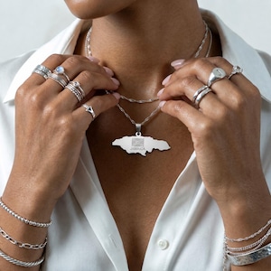 May include: A silver necklace with a pendant in the shape of Jamaica, featuring the Jamaican flag. The image also shows various silver rings and bracelets. The word "JAMAICA" is visible on the pendant.
