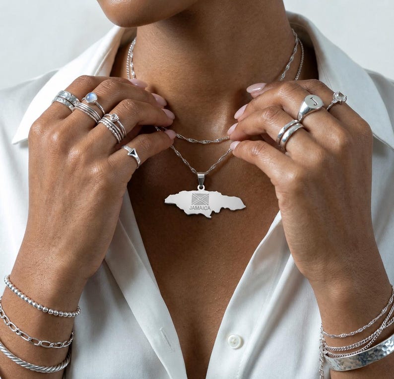 May include: A silver necklace with a pendant in the shape of Jamaica, featuring the Jamaican flag. The image also shows various silver rings and bracelets. The word "JAMAICA" is visible on the pendant.