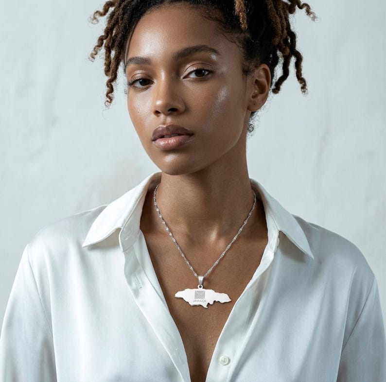 May include: A silver necklace featuring a pendant in the shape of Jamaica. The pendant has a detailed design of the Jamaican flag. The necklace is worn by a person wearing a white button-down shirt.