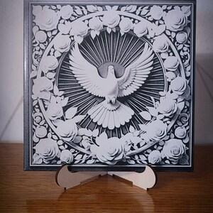 Op de afbeelding: Vierkant zwart-wit kunstwerk met een duif met uitgespreide vleugels, omringd door een bloemenkrans. Het kunstwerk wordt gepresenteerd op een houten standaard, met een gedetailleerd grijstinten ontwerp.