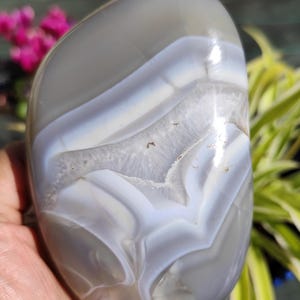 May include: A polished, oval-shaped agate stone. The stone features bands of gray and white, with a translucent quality. The natural patterns create a unique, swirling design. The stone is held in a hand, with greenery and pink flowers in the background.