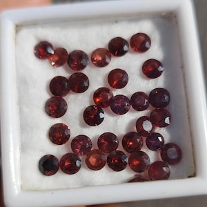 Puede incluir: Una colección de gemas redondas, facetadas, de color rojo oscuro, presentadas en un recipiente cuadrado blanco. Las gemas tienen una superficie pulida y reflectante, y están dispuestas muy juntas. El recipiente está lleno de un material blanco y suave.