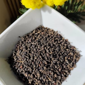 Pode incluir: Uma tigela quadrada branca cheia de bagas secas castanhas escuras. As bagas são pequenas e agrupadas. Flores amarelas brilhantes no fundo, adicionando um toque de cor. A imagem é bem iluminada, destacando a textura das bagas.