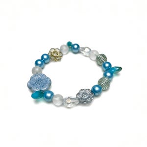 May include: A beaded bracelet featuring a mix of blue, silver, and clear beads. The bracelet includes round beads, flower-shaped beads, and other decorative elements. The color palette is primarily cool tones, with a touch of gold.