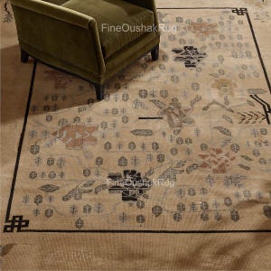 Peut inclure: Tapis Oushak beige avec une bordure noire et des motifs floraux et géométriques complexes dans des tons de gris, marron et orange. Un fauteuil vert est dans le coin supérieur gauche. Le tapis a le texte "FineOushakRug".