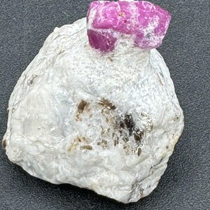 Puede incluir: Una gema de rubí en bruto y sin cortar, de un color rojo rosado vibrante, se asienta sobre una roca blanca y texturizada. La superficie del rubí tiene una apariencia cristalina. La roca tiene una superficie rugosa e irregular con algunas inclusiones marrones y beige.