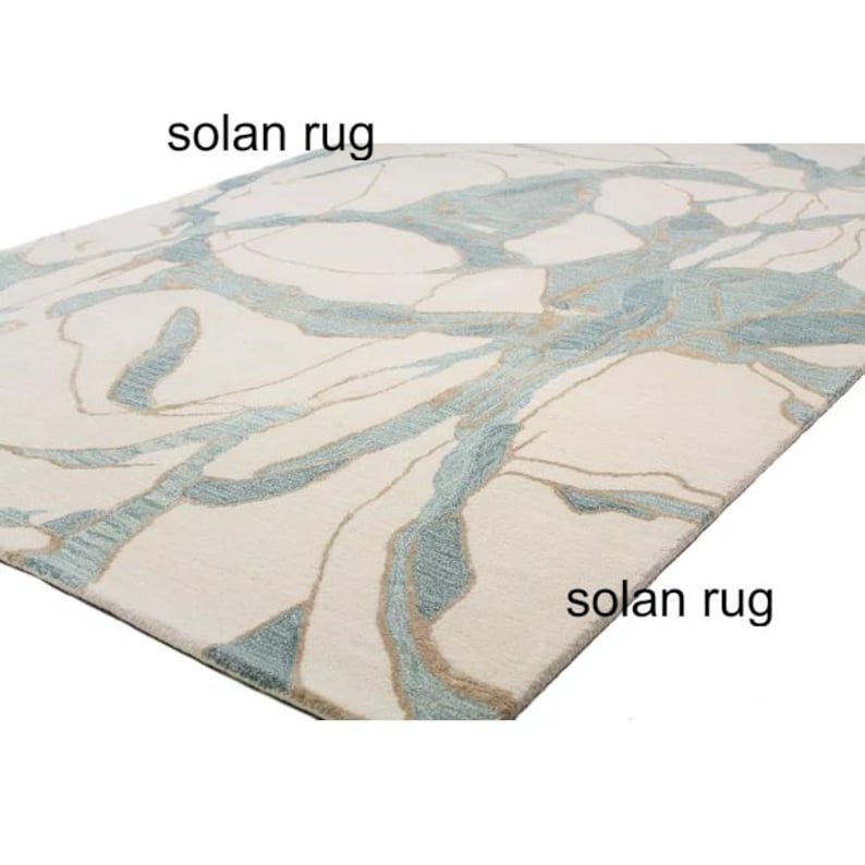 May include: A rectangular rug with a cream-colored background and a pattern of blue and tan abstract lines. The text "solan rug" is printed on the rug.