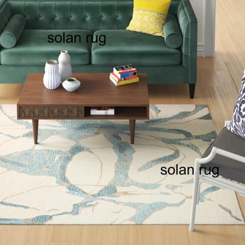 May include: A living room scene featuring a teal sofa, a wooden coffee table, and a patterned rug. The rug has a cream background with blue and gold abstract designs. Decorative vases and books are on the table. The text "solan rug" is visible.